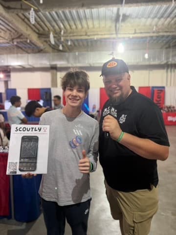 Henry holding a Scoutly flyer while speaking with a FUN Robotics Network interviewer at a robotics event.